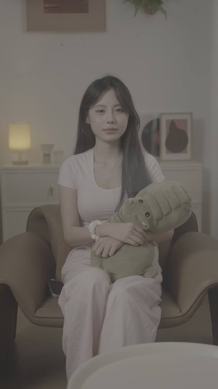 原始实拍 · 粉衣人物抱绿色玩偶的正面口播原始帧。